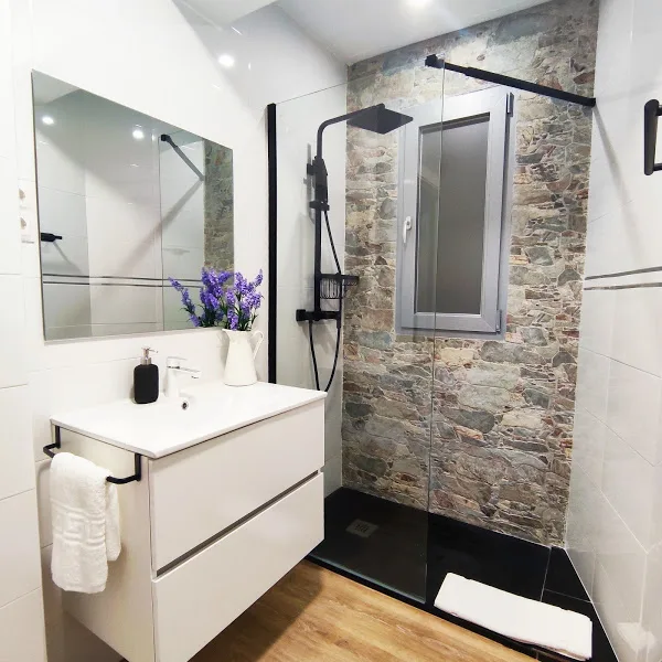 Baño moderno con ducha tipo lluvia, pared de piedra, espejo amplio y muebles blancos en apartamento turístico en Galicia