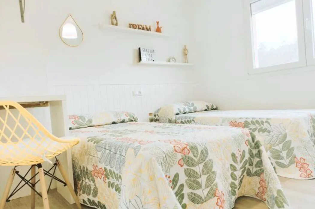 Dormitorio luminoso con dos camas individuales, colchas florales, decoración minimalista y ventana con vistas en alojamiento turístico en Galicia